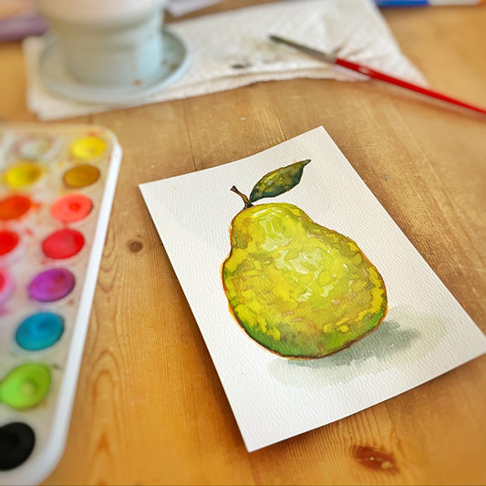 A watercolor painted pear on a small white card placed on a wooden table, with a paint palette, brush, and paint container blurred in the background.