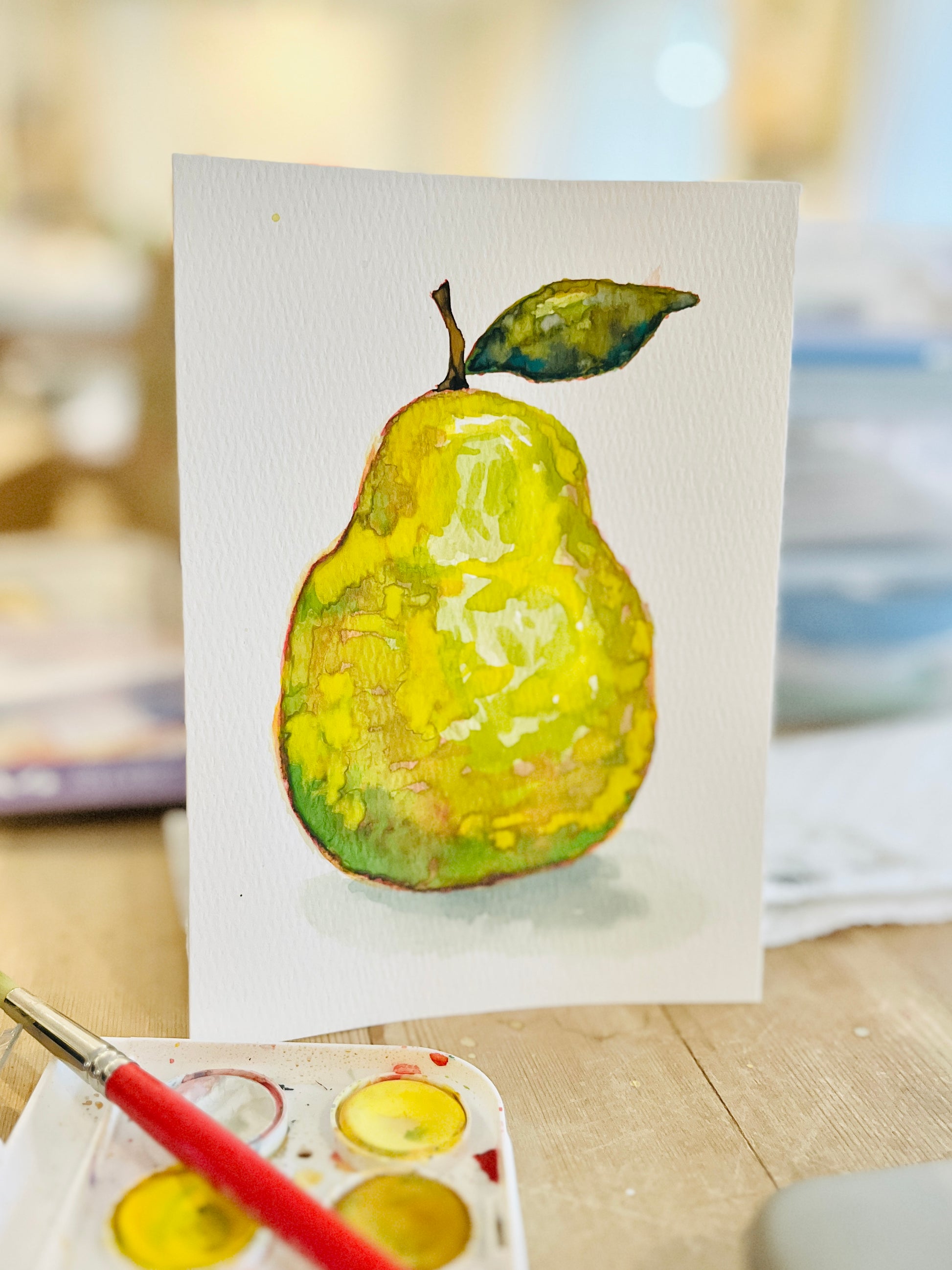 A vertical watercolor painting of a yellow pear with a green leaf on a white card, displayed on a wooden surface with a paint palette and brush in the foreground.