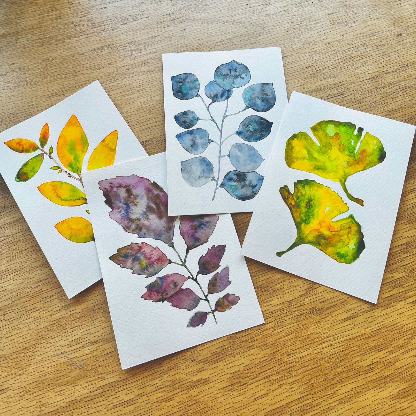 Four small art prints featuring watercolor illustrations of leaves in yellow, blue, and purple tones, arranged on a light wood tabletop.