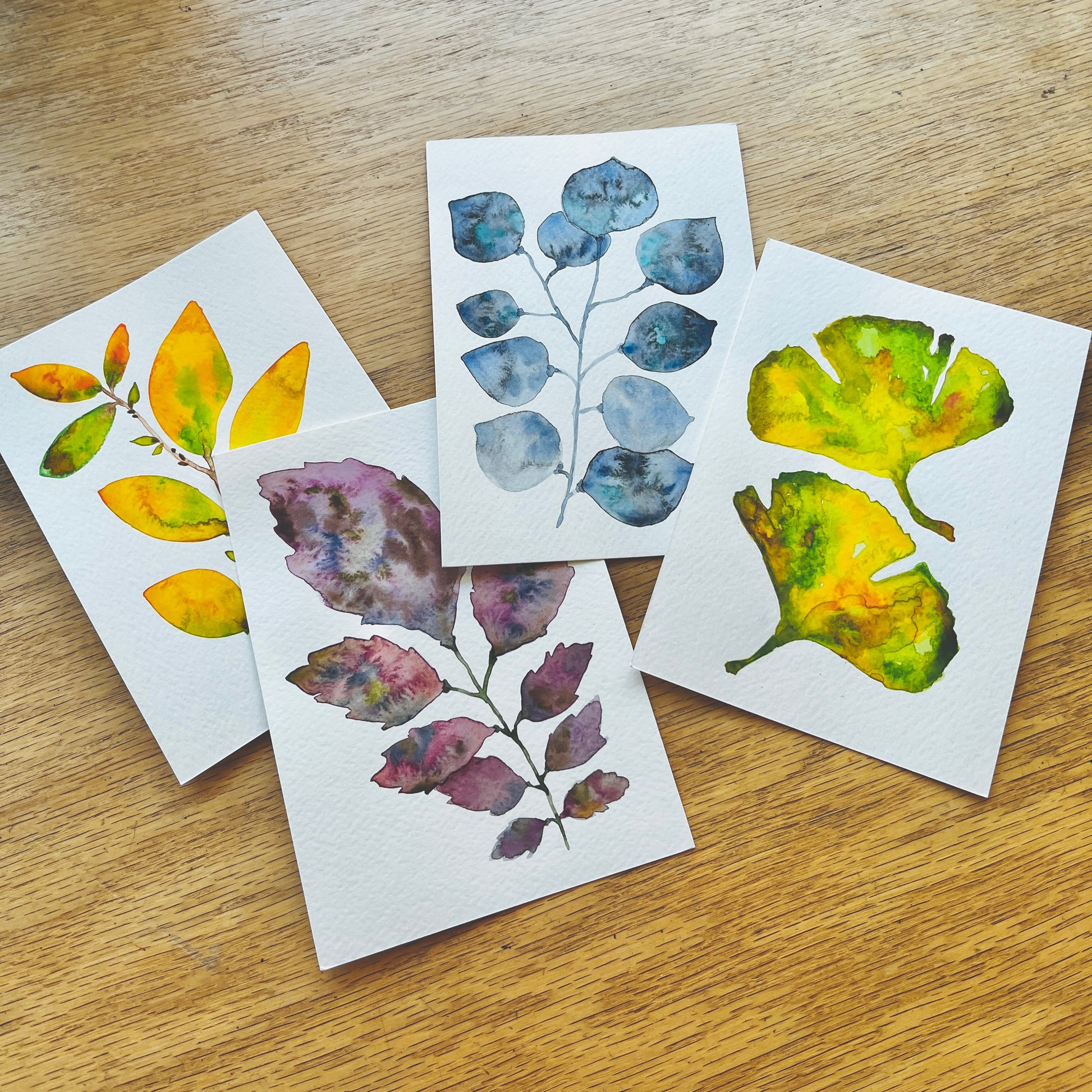 Four small art prints featuring watercolor illustrations of leaves in yellow, blue, and purple tones, arranged on a light wood tabletop.