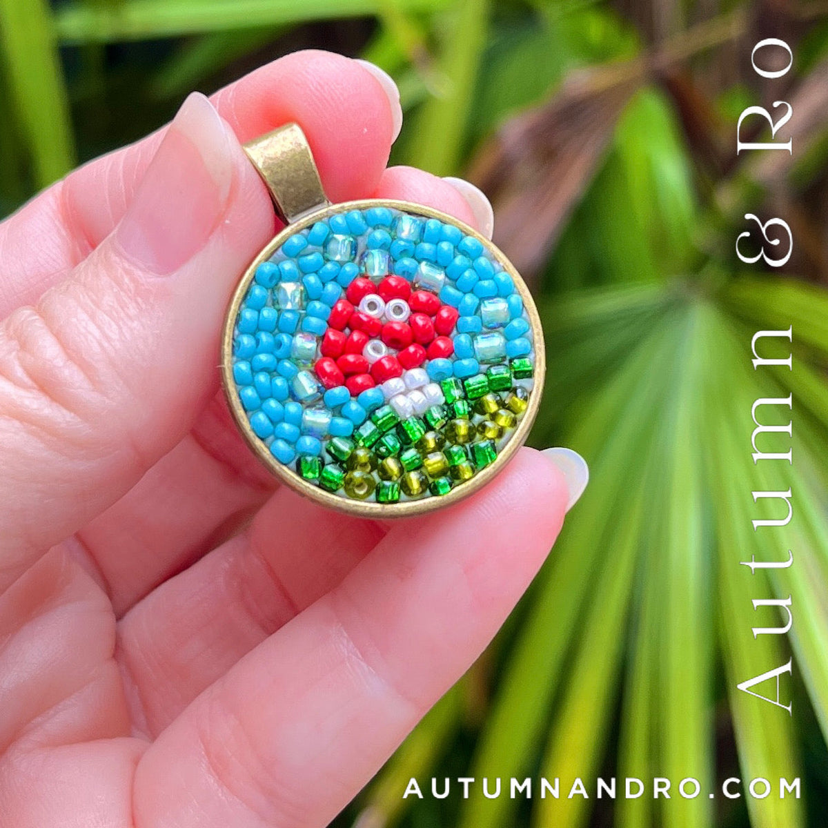 A hand holds a round mosaic beaded pendant featuring a red mushroom on a blue background with green and gold details, set against a blurred green foliage background. 